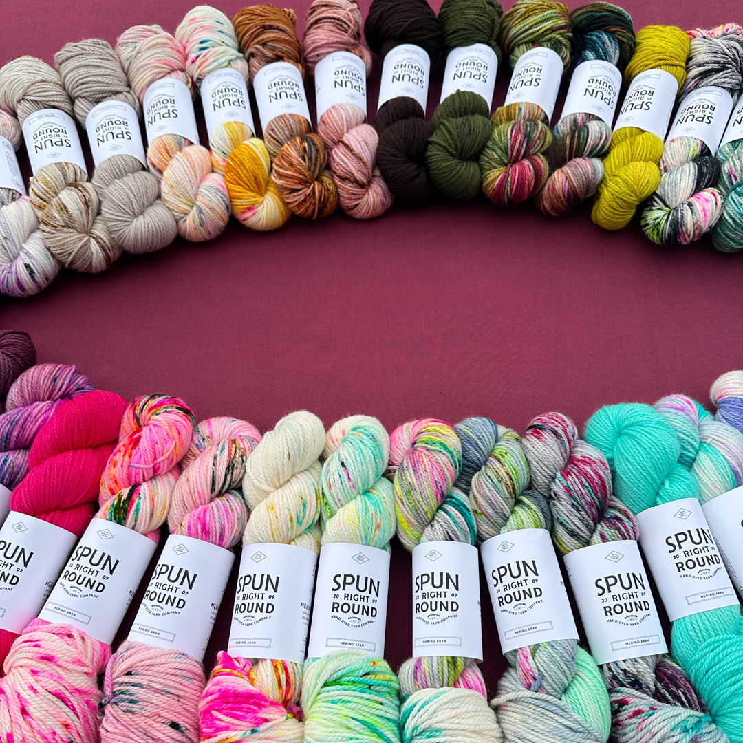 Skeins of Spun Right Round hand-dyed yarn arranged in a semi-circle against a solid magenta background. The yarn displays a vibrant spectrum of colors, including hot pink, neon orange, speckled cream, mint green, and deep teal. Each skein is wrapped in a clean white paper label with the brand’s logo and name printed in bold, black typography.