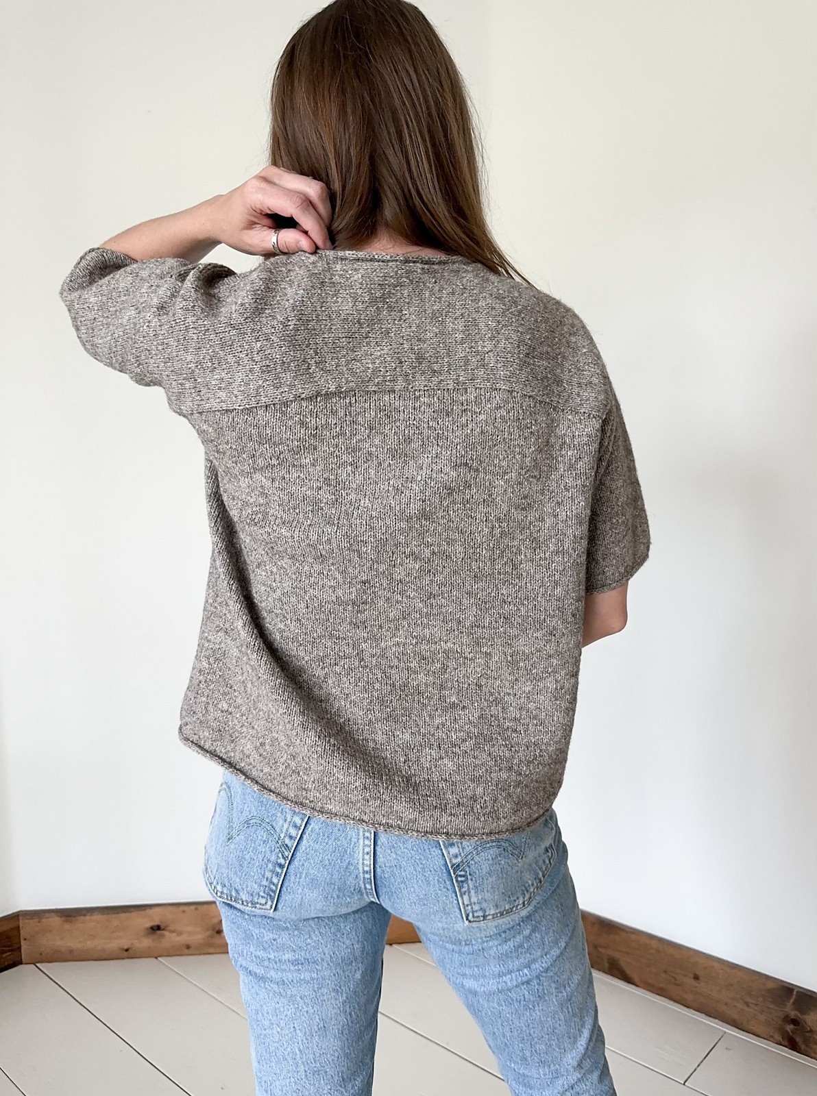 Person with long hair wearing a textured gray/beige short-sleeve knit sweater and blue jeans, standing against a plain light gray wall.