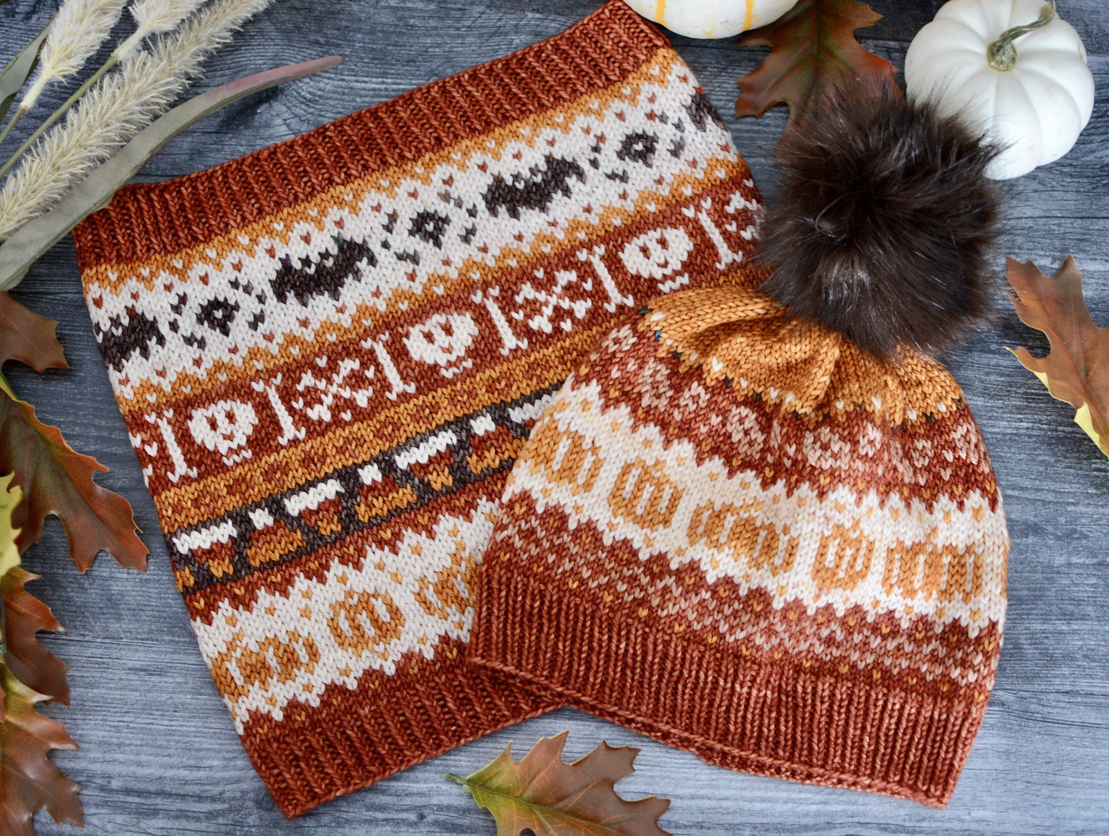 The Autumn Doodle knitted hat and cowl (cowl pattern sold separately). The cowl features pumpkins, candy corn, skulls and bones, and bats in shades of orange, rust, brown, and cream yarn. The hat features pumpkins and fair-isle style diamonds in shades of orange, rust, and cream, and includes a fuzzy faux-fur pompom on top. Autumn leaves and white pumpkins decorate the gray background. 