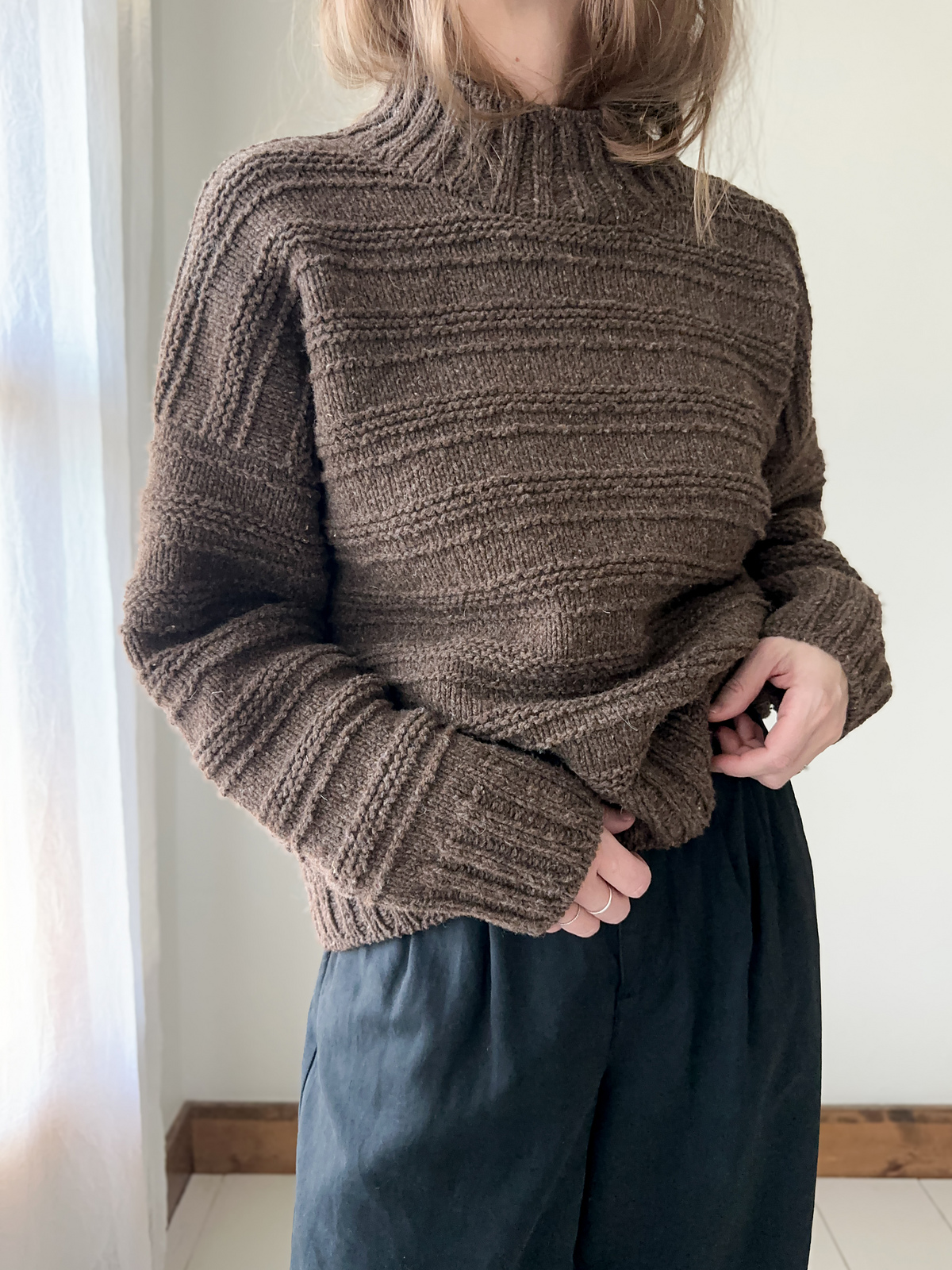 A person with light skin and short blond hair models a textured brown pullover sweater. The sweater features a wide-ribbed funnel neck and matching ribbing on cuffs and hem, and rows of garter stitch interspersed with wider rows of stockinette.