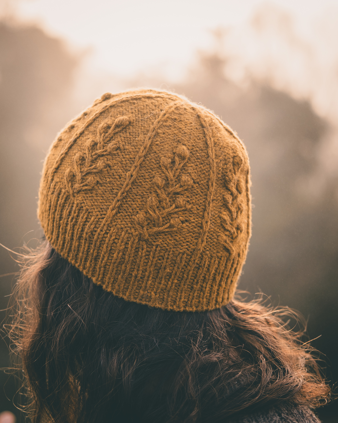 A person with long brown hair wearing a hand knitted dark yellow hat faces towards a background of trees seen through a hazy golden mist. The hat features a botanical motif created from bobbles and cables, resting on top of rows of purl stitches. The brim has a 1 x 1 rib.
