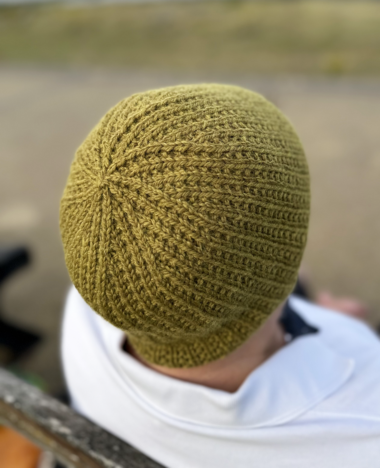 A person in a white sweatshirt faces away from us, modeling a textured beanie hat knitted in deep gold yarn.