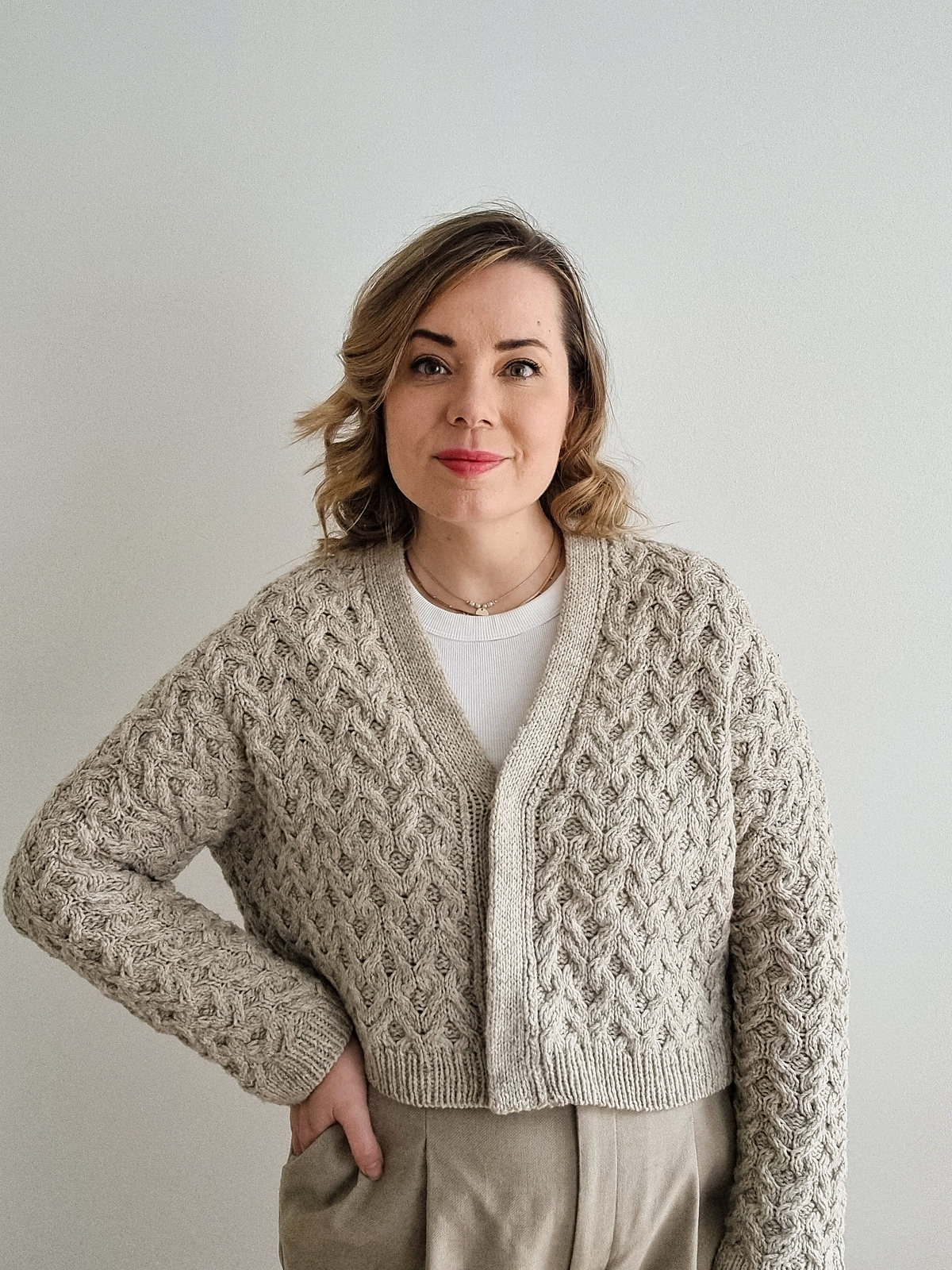 Knitting designer Sari Nordlund, a white woman with dark blonde shoulder-length hair, models a hand knitted cabled cardigan in pale gray wool yarn. The cardigan is slightly cropped and has a v-neck with a simple button band and ribbing at hem and cuffs.