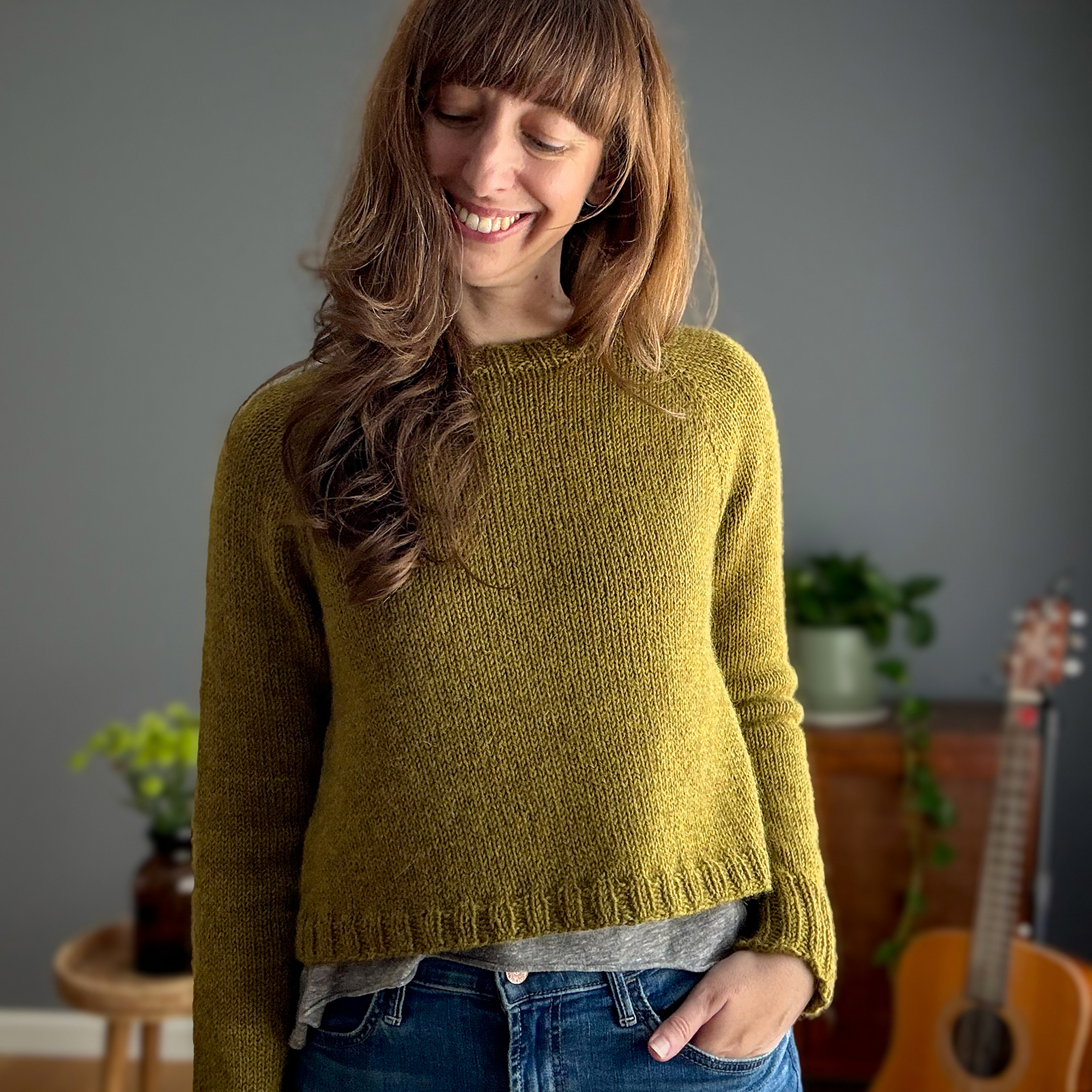 A smiling white woman with long brown hair models a hand knitted pullover sweater in deep yellow-green wool yarn.