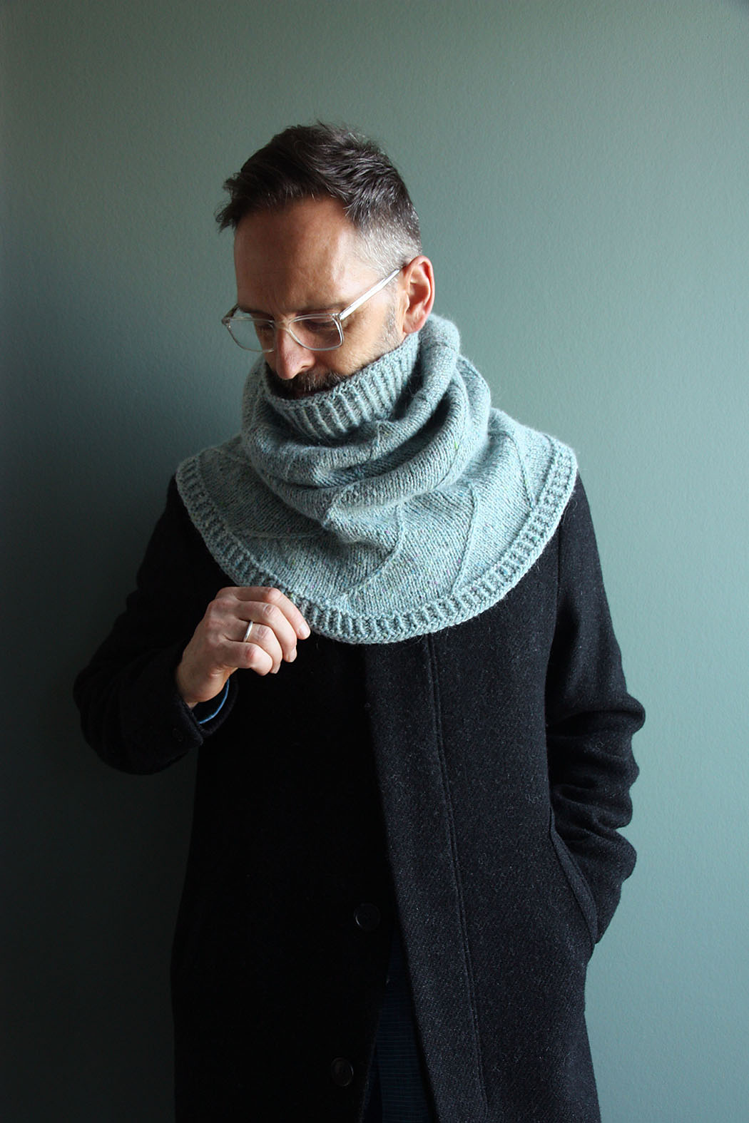 A white man with short dark hair and glasses models a knitted cabled cowl in pale blue-green wool yarn.