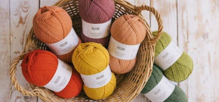 Several skeins of wool yarn in fall colors of red, orange, and gold lie in a wicker basket, with two more skeins in shades of green resting nearby on the whitewashed wooden floor