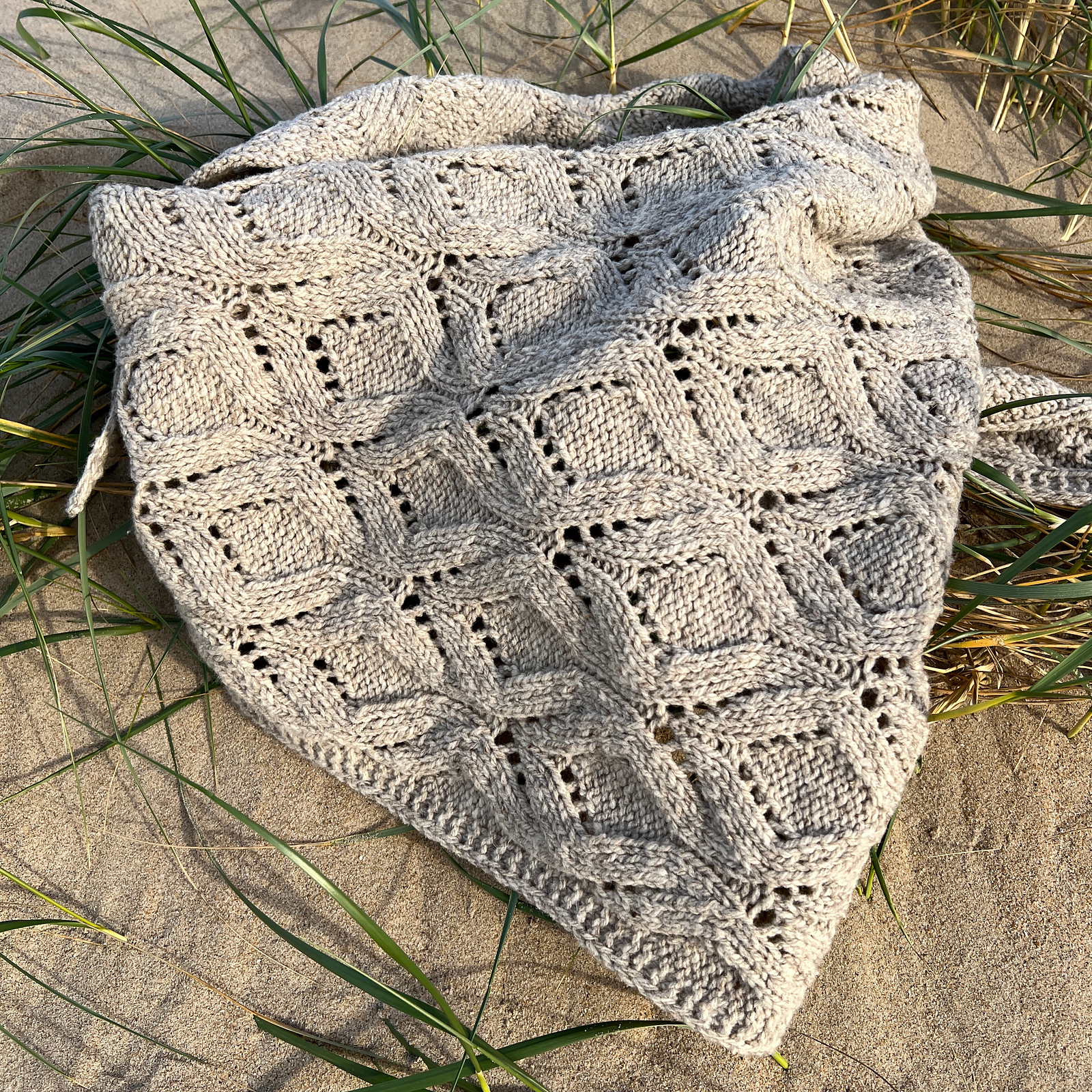 A pale gray handknitted lace shawl lies on beach grasses, lit by warm golden sunlight. The wavy cables create soft diamond shapes highlighted by the lacework.