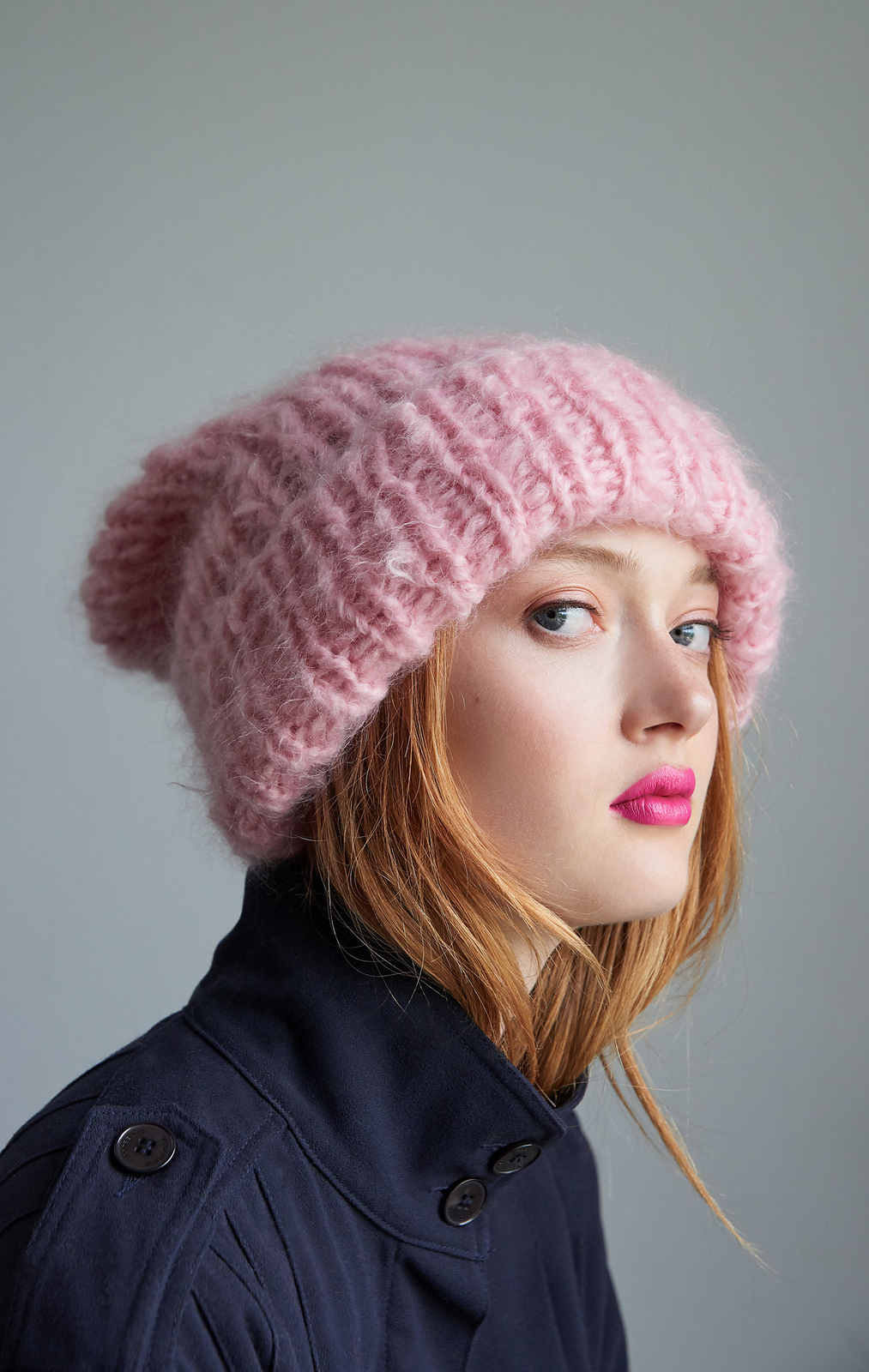 A young white woman with long red hair models a fluffy ribbed hat knitted in pink mohair yarn.