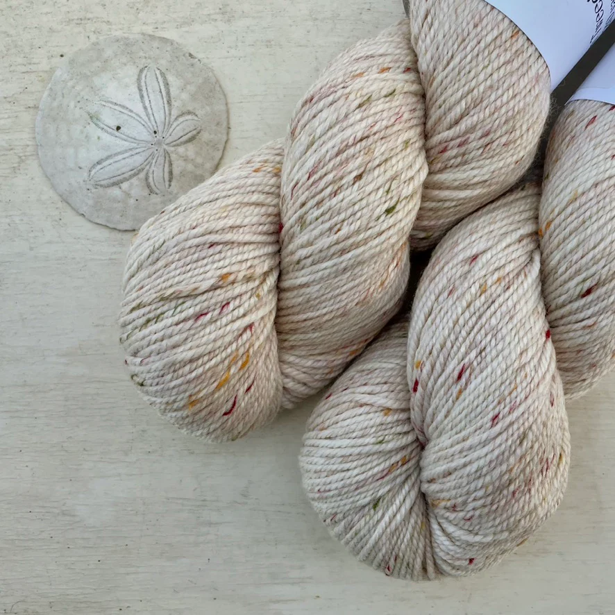 Two skeins of pale beige tweedy wool yarn lie next to a sand dollar. 