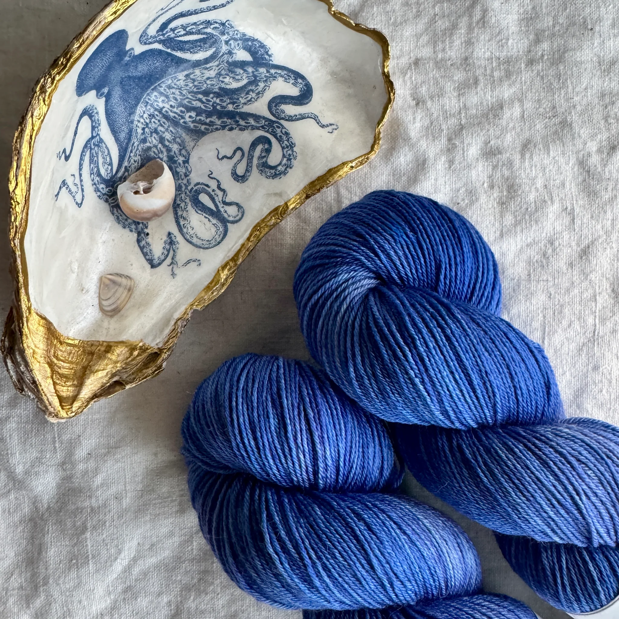 Closeup of two skeins of vibrant blue yarn next to a large gilded seashell painted with a blue octopus that holds smaller seashell.