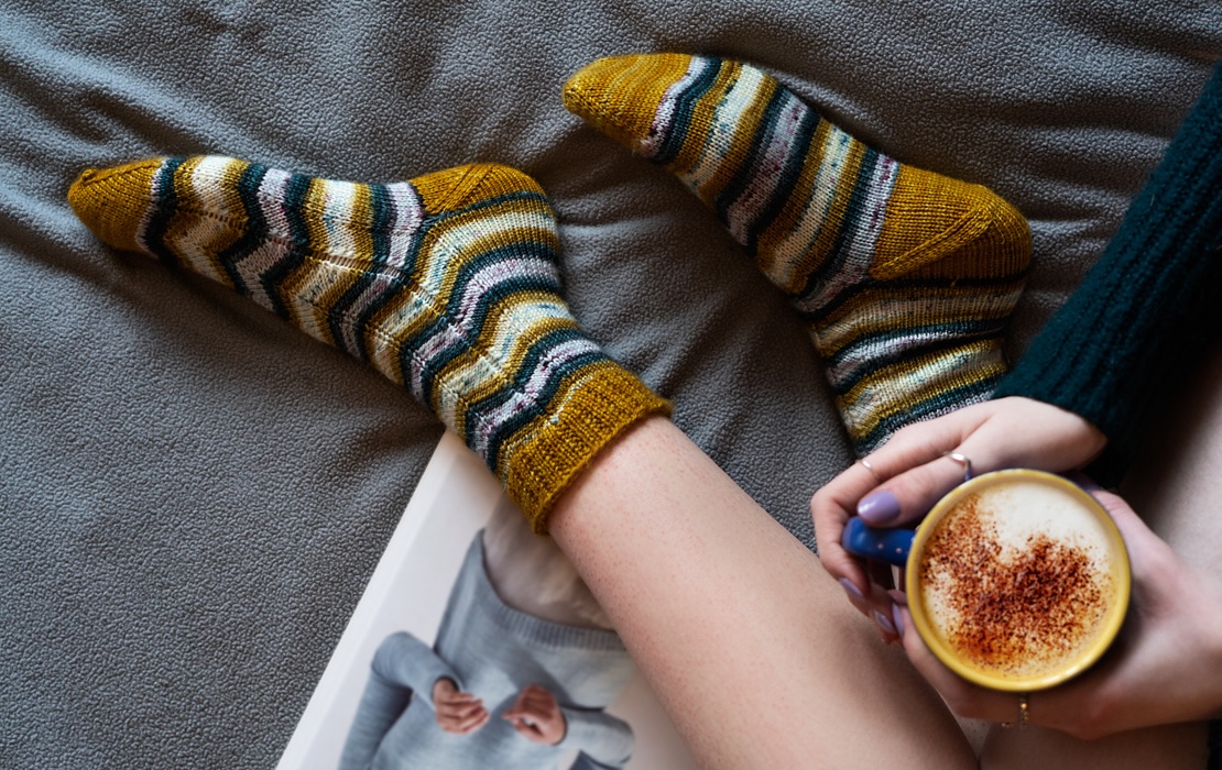 Closeup of a person's feet in chevron-striped hand knitted socks in shades of gold and blue.
