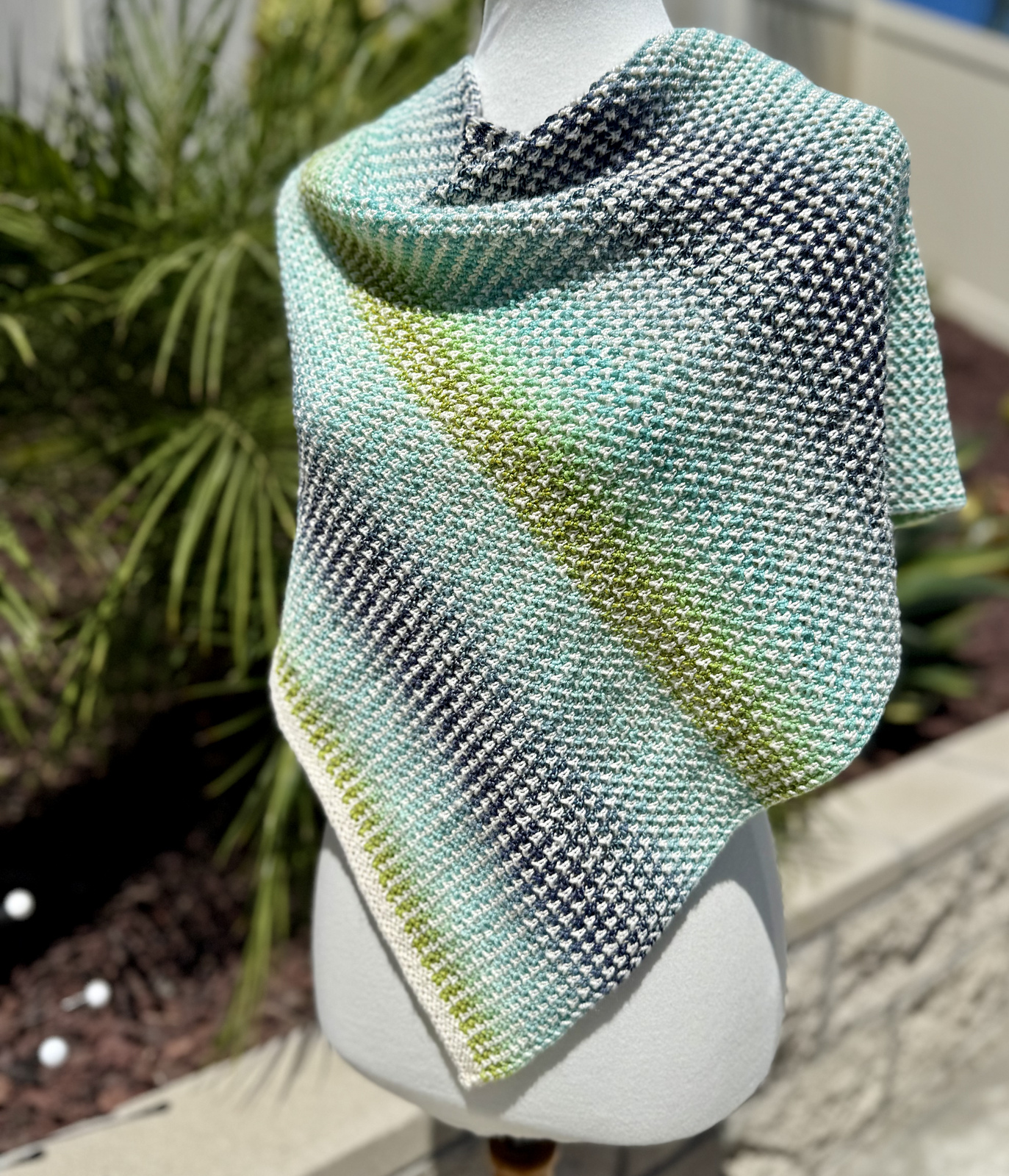 A hand knitted shawl striped in shifting shades of blues and greens is draped over a dress form.