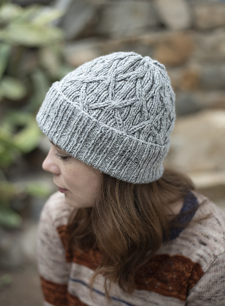 A white woman with long brown hair models a cabled hat knitted in pale gray yarn. 