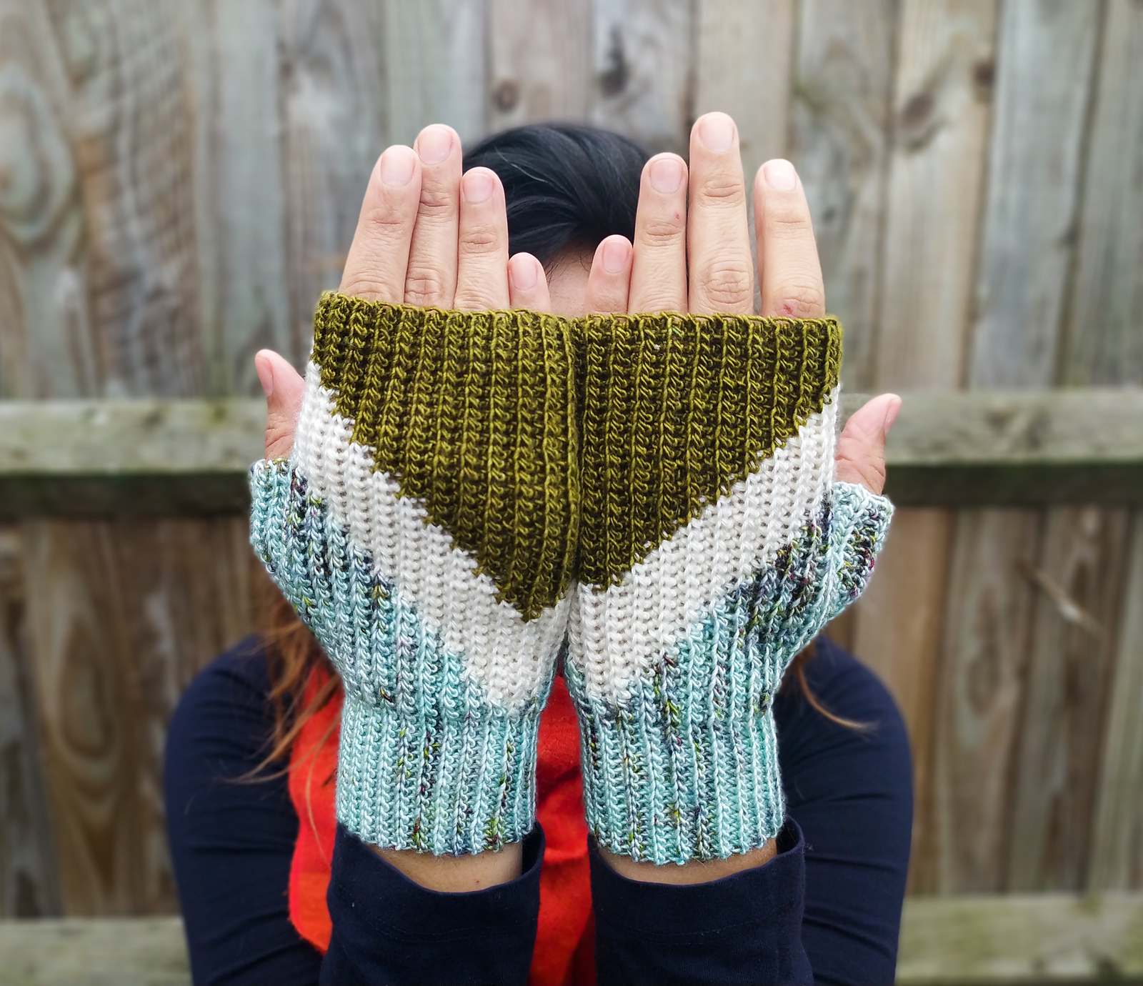 A light-skinned person holds their hands up in front of their face to show off their diagonally striped crocheted mitts.