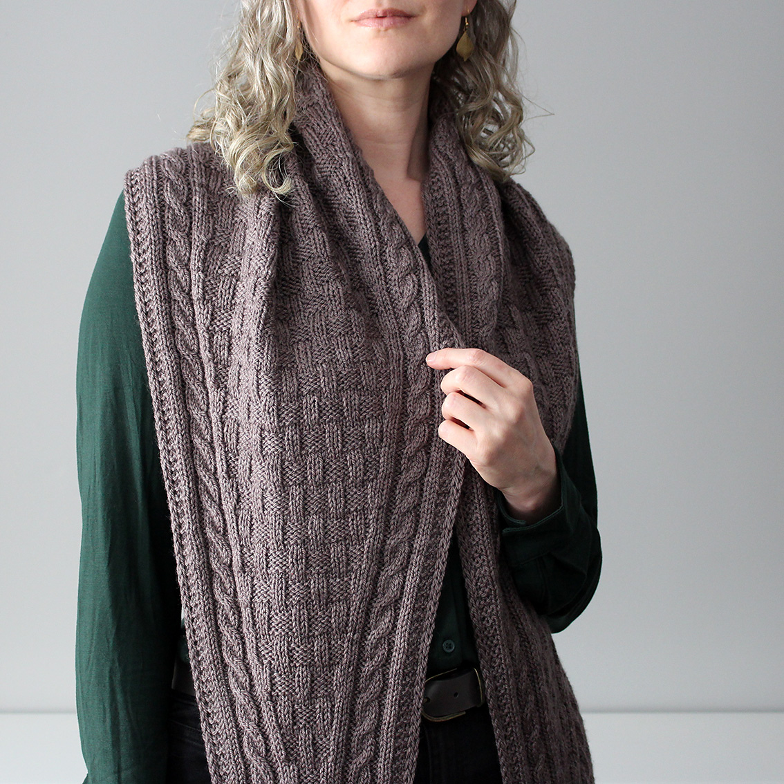 A white woman with wavy blonde hair models a knitted wrap with a checkerboard texture and cabled borders in soft brown.