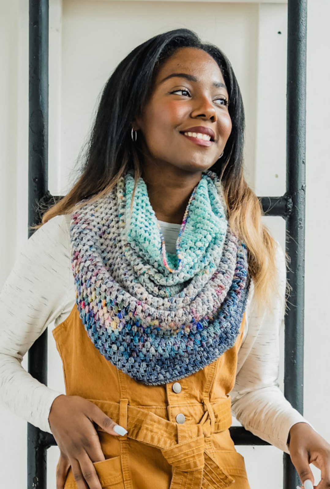 A brown-skinned woman with long dark hair wears a crocheted cowl in shades of blue over bright yellow-orange overalls.