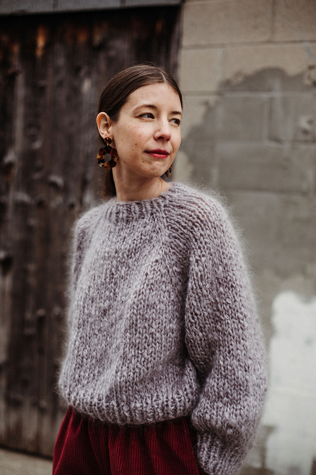 A white woman with straight brown hair models a fuzzy loosely knit pullover in soft lavender mohair yarn. The pattern has a crew neck, raglan shaping, and slightly billowy sleeves that are cuffed at the wrists.