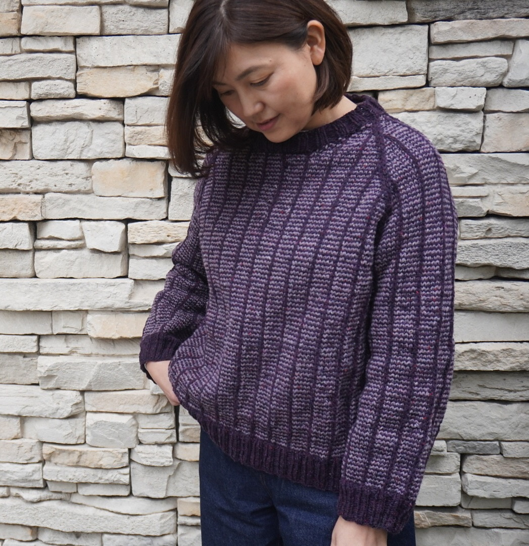 A Japanese woman with short dark hair examines her sweater, knitted in a laddered striped pattern of light and dark purple yarns.
