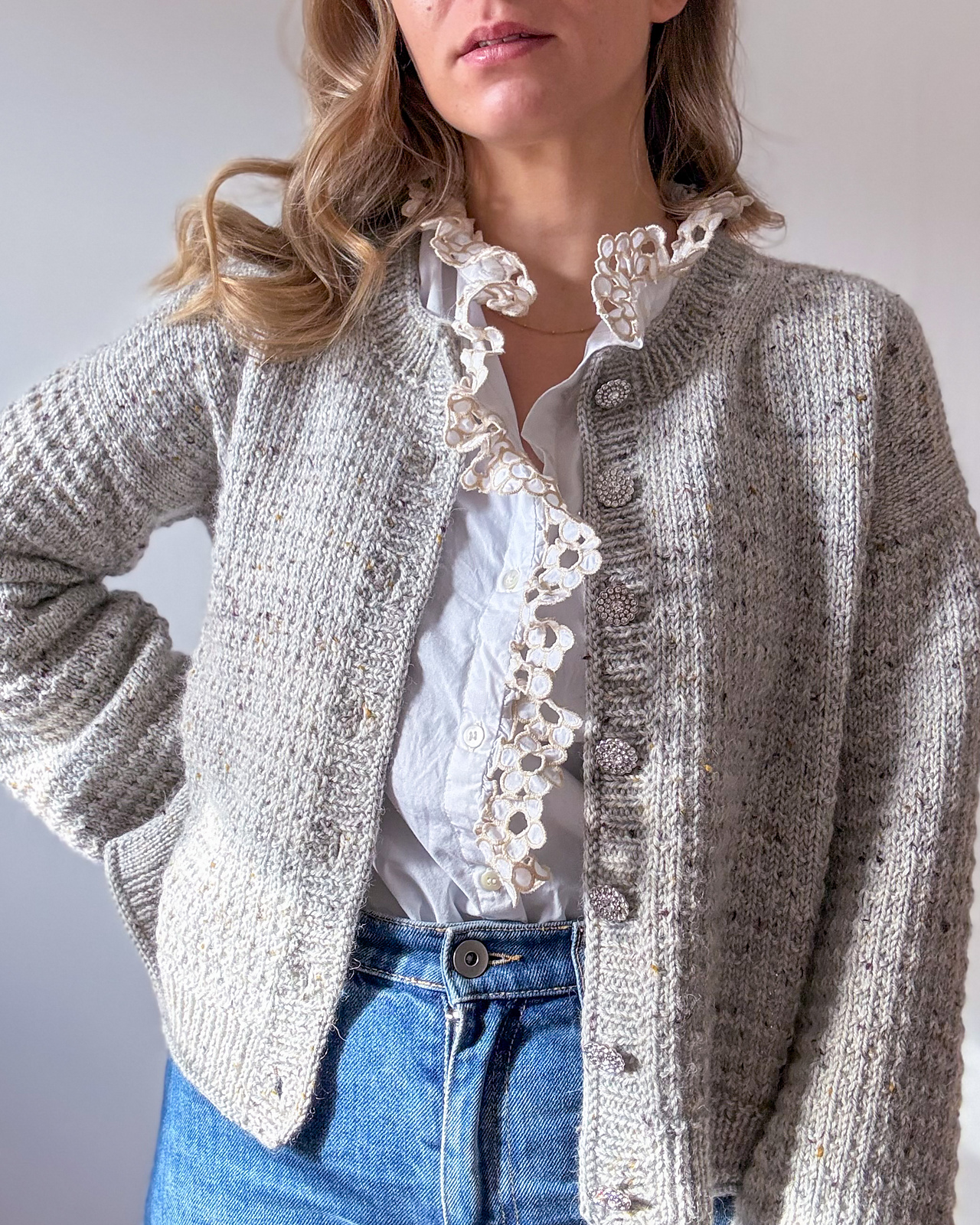 A white woman with long blonde hair wears a hand knitted cardigan in pale gray wool over a ruffled white blouse and blue jeans.