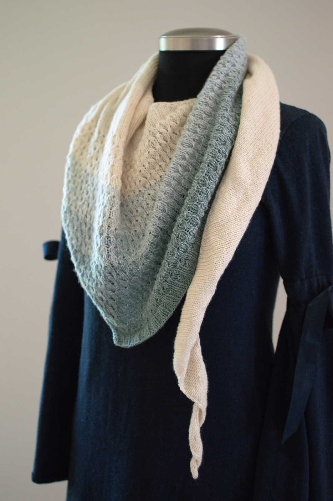 A textured shawl in a gradient of soft blues and sandy beige colors, knitted in a drapey textured stitch pattern.
