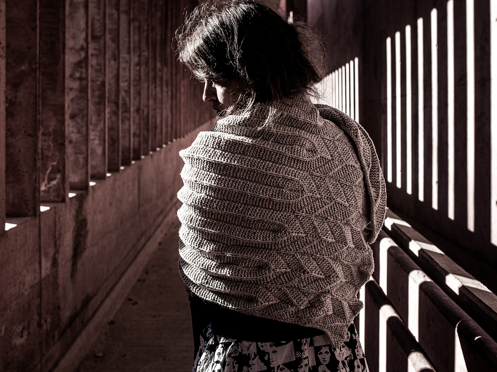 A dark haired person wears a deeply textured shawl knitted in dark gray-brown wool. They are facing away from the camera and are dramatically lit by rays of sunlight slanting through concrete slats.