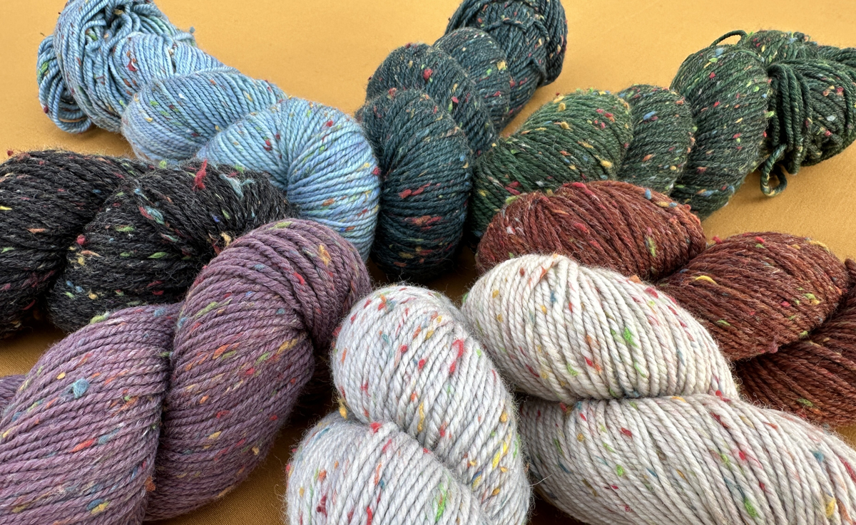 A circle of tweedy wool yarn skeins in beige, orange, green, blue, and purple, on a warm yellow background.