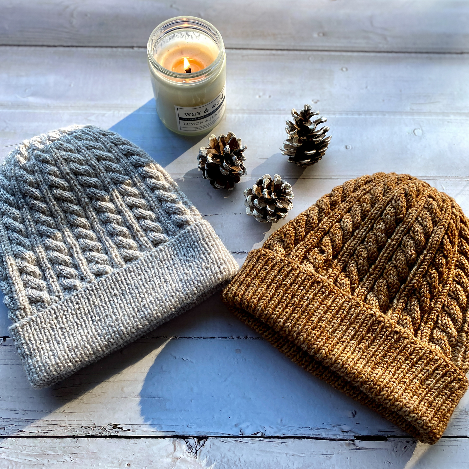 Two cabled hats knitted in soft gray and golden brown lie on a white wooden table next to a lit candle and three pinecones.