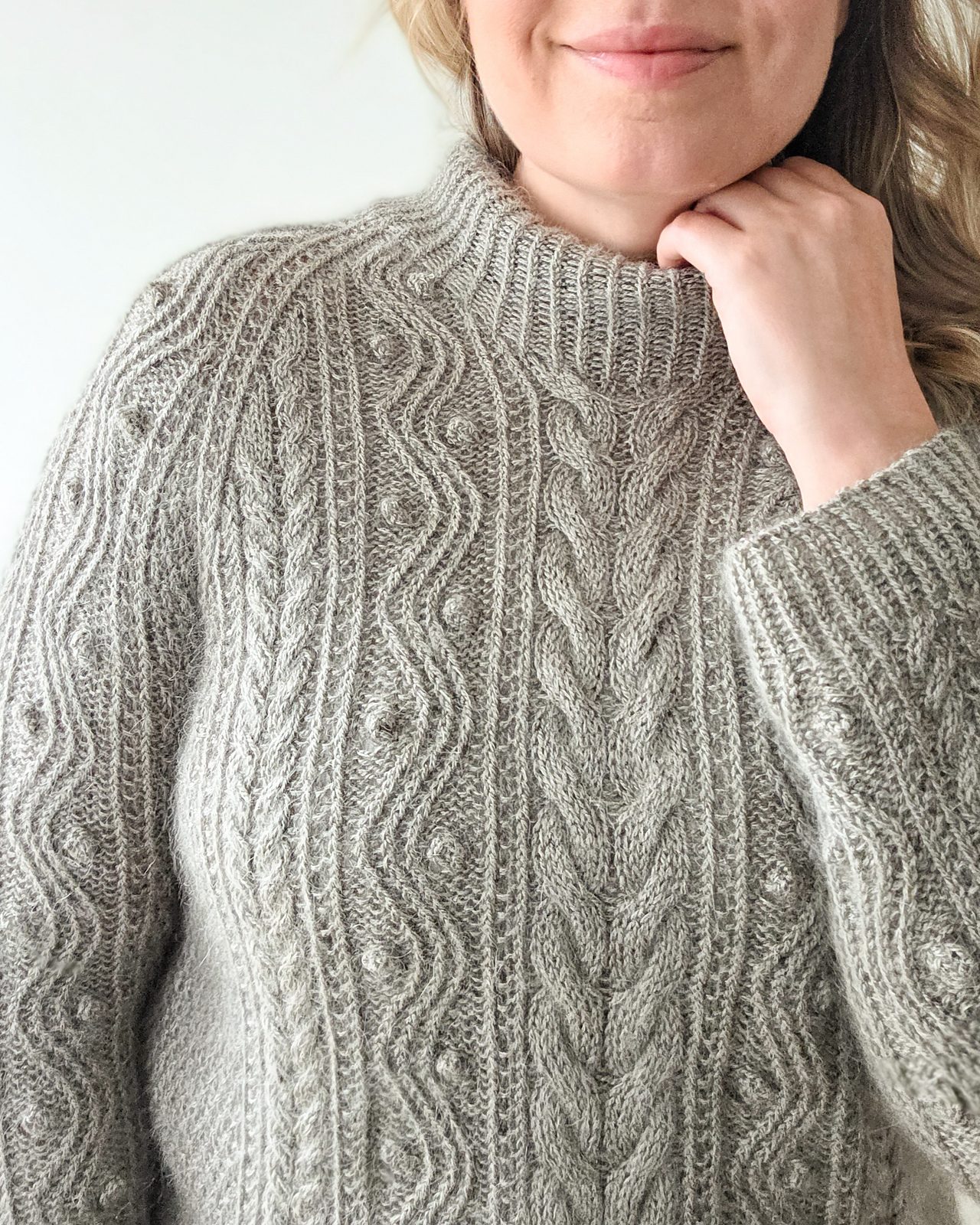 Closeup of a white woman with long dark blonde hair wearing a hand knitted cabled pullover in pale gray wool yarn.