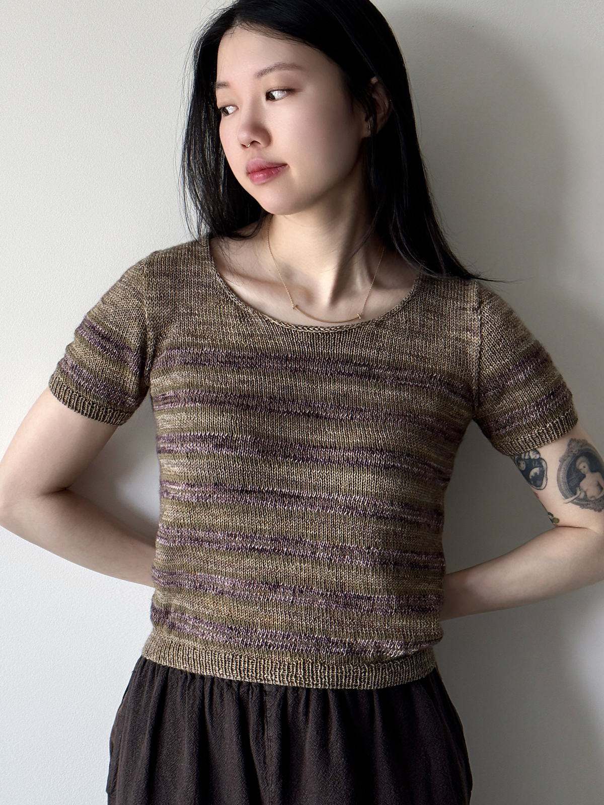 A light-skinned woman with long dark hair models a short sleeved striped sweater knitted in shades of deep brown and plum.