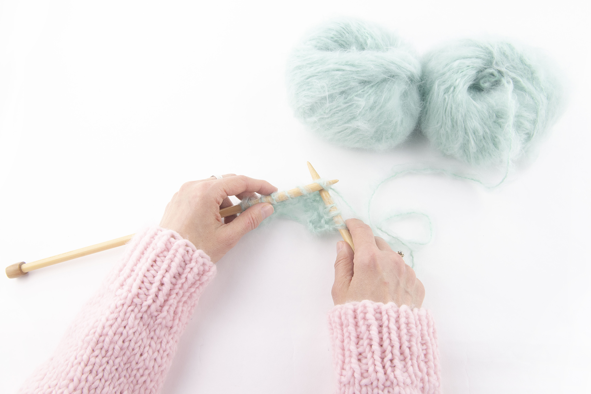A lightskinned person's hands hold wooden knitting needles. They wear a fluffy pink ribbed sweater and are knitting with fluffy aqua colored yarn.