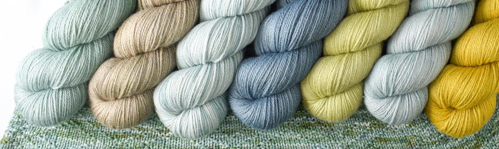 Several skeins of silky yarn in soft shades of blue, green and yellow lie side by side on a soft green knitted shawl.