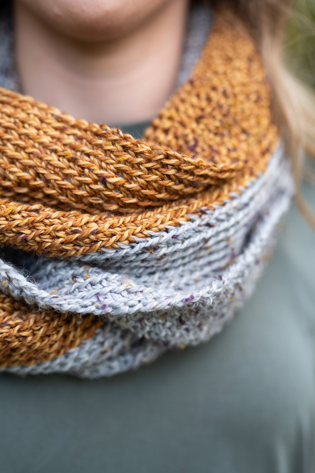 Closeup of a light-skinned person wearing a crocheted cowl in pale gray and deep gold wool yarn.