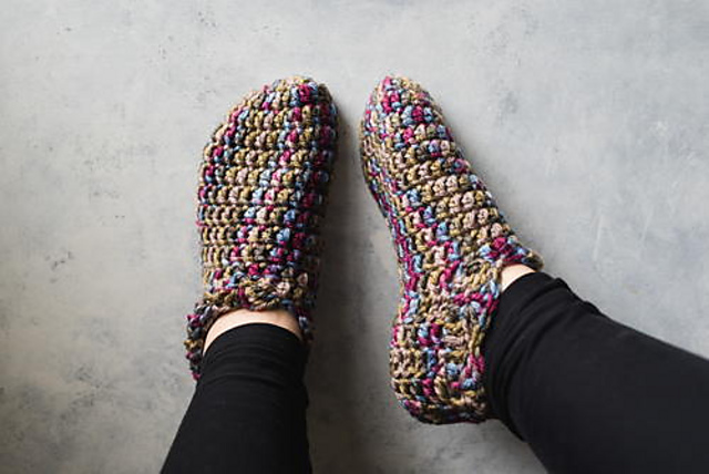 Two feet wearing colorful crocheted slippers resting on a light grey surface. The slippers feature a mix of earth tones and vibrant pinks, blues, and greens.