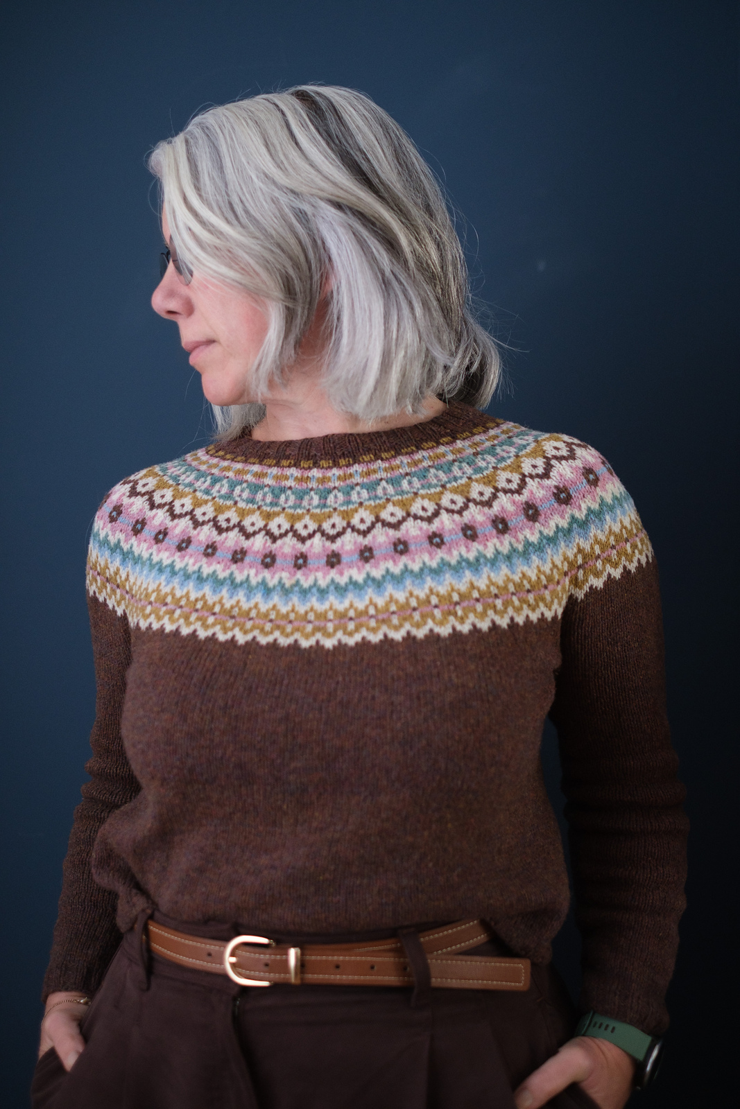 A white woman with shoulder length gray hair models a knitted pullover in warm brown wool with a colorwork yoke in white, gold, pink and blue.