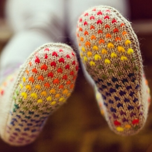 Close-up of colorful, knitted socks showing soles with vibrant dots in a rainbow pattern against a blurred background. The socks convey warmth and comfort.