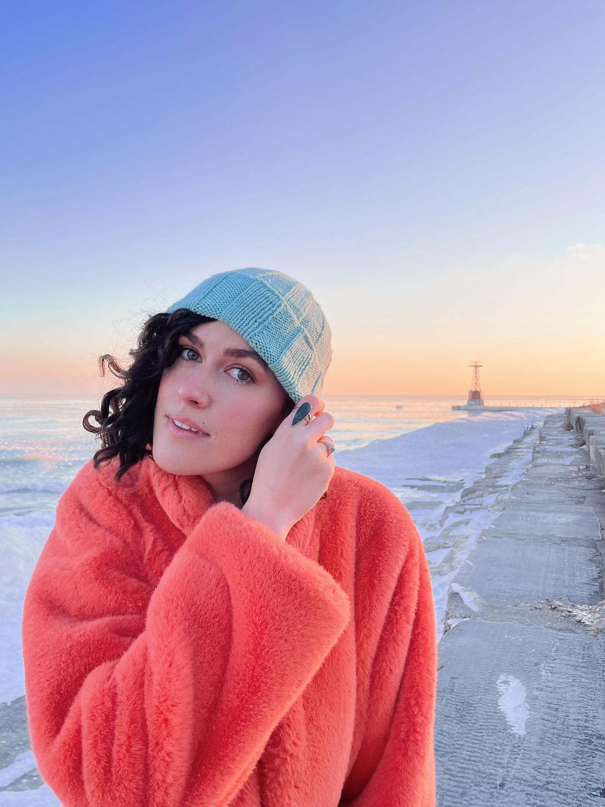 Knitwear designer Park Williams, a white woman with wavy dark hair, models a knitted ribbed beanie in pale blue yarn along with a bright fuzzy coat.