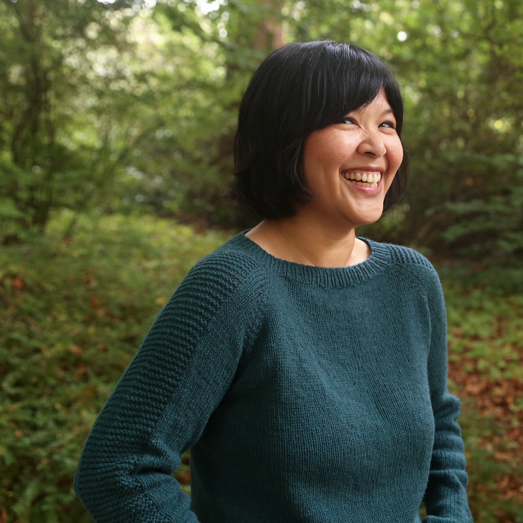 A smiling Asian woman with short dark hair stands in a green forest wearing a knitted pullover in deep teal yarn