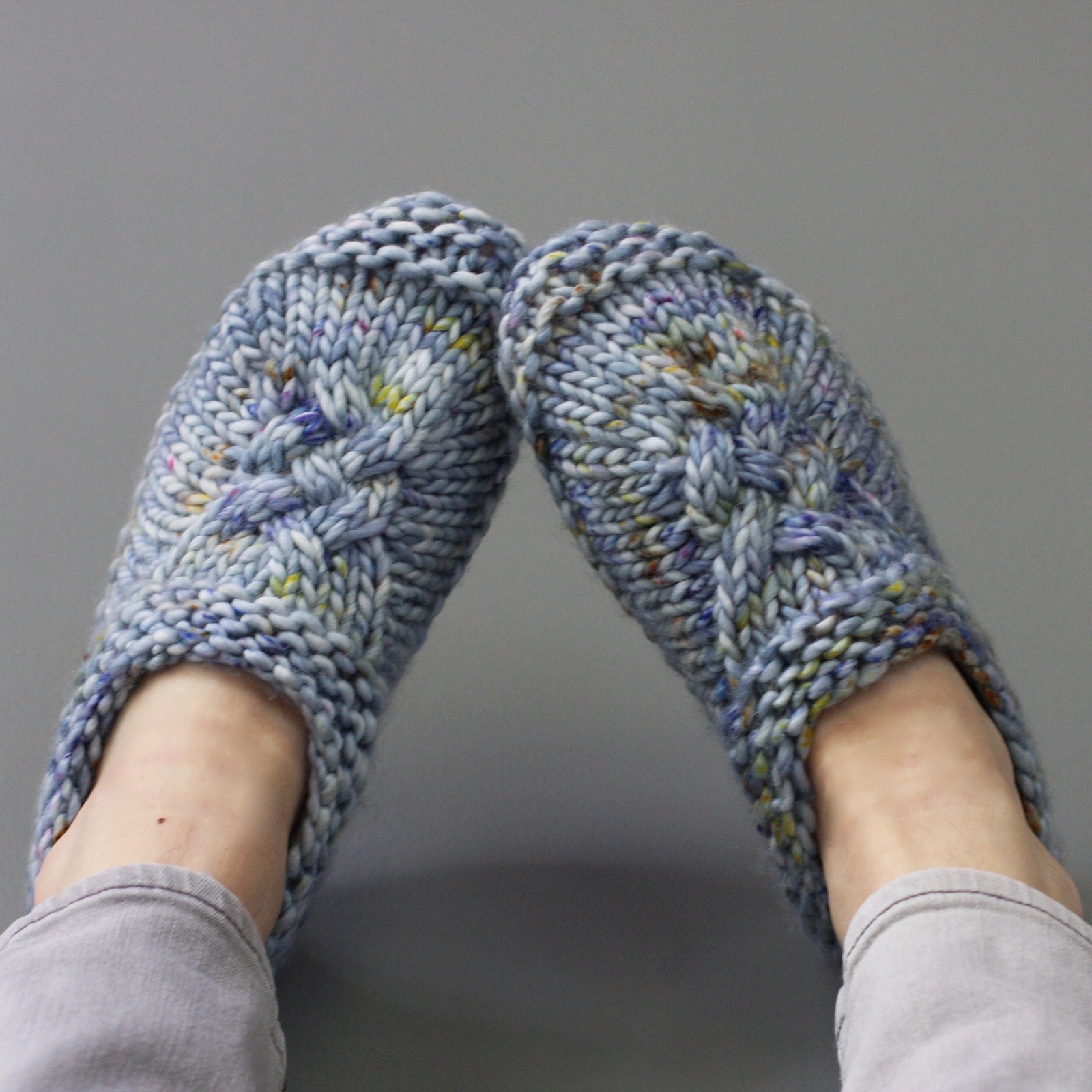 Feet wearing cozy, hand-knitted blue slippers with a textured pattern in a relaxed pose. The slippers feature multicolored speckles, conveying warmth.