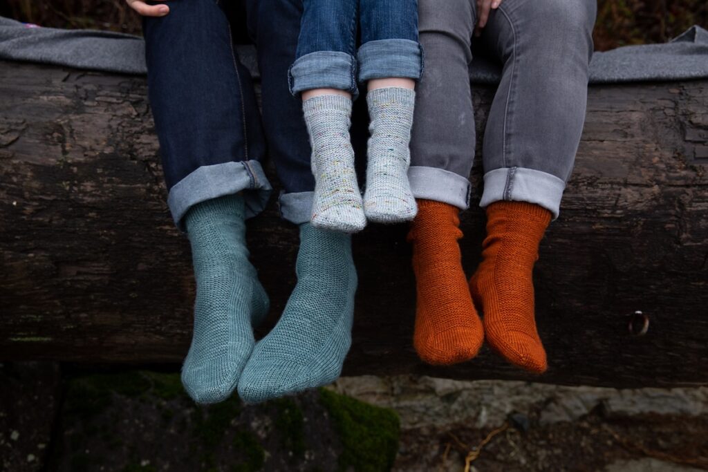 Closeup of the legs and feet of two adults and a child wearing hand knitted socks in soft teal, pale gray, and deep orange.
