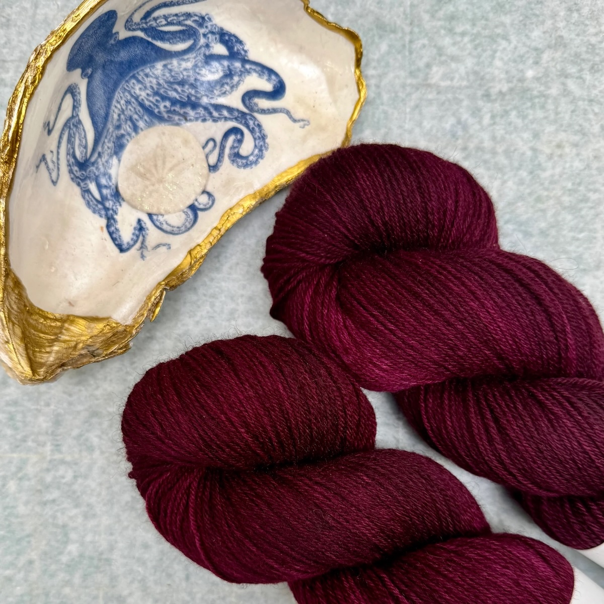 Closeup of two skeins of deep berry red yarn lying next to a gold-edged seashell painted with a blue octopus.