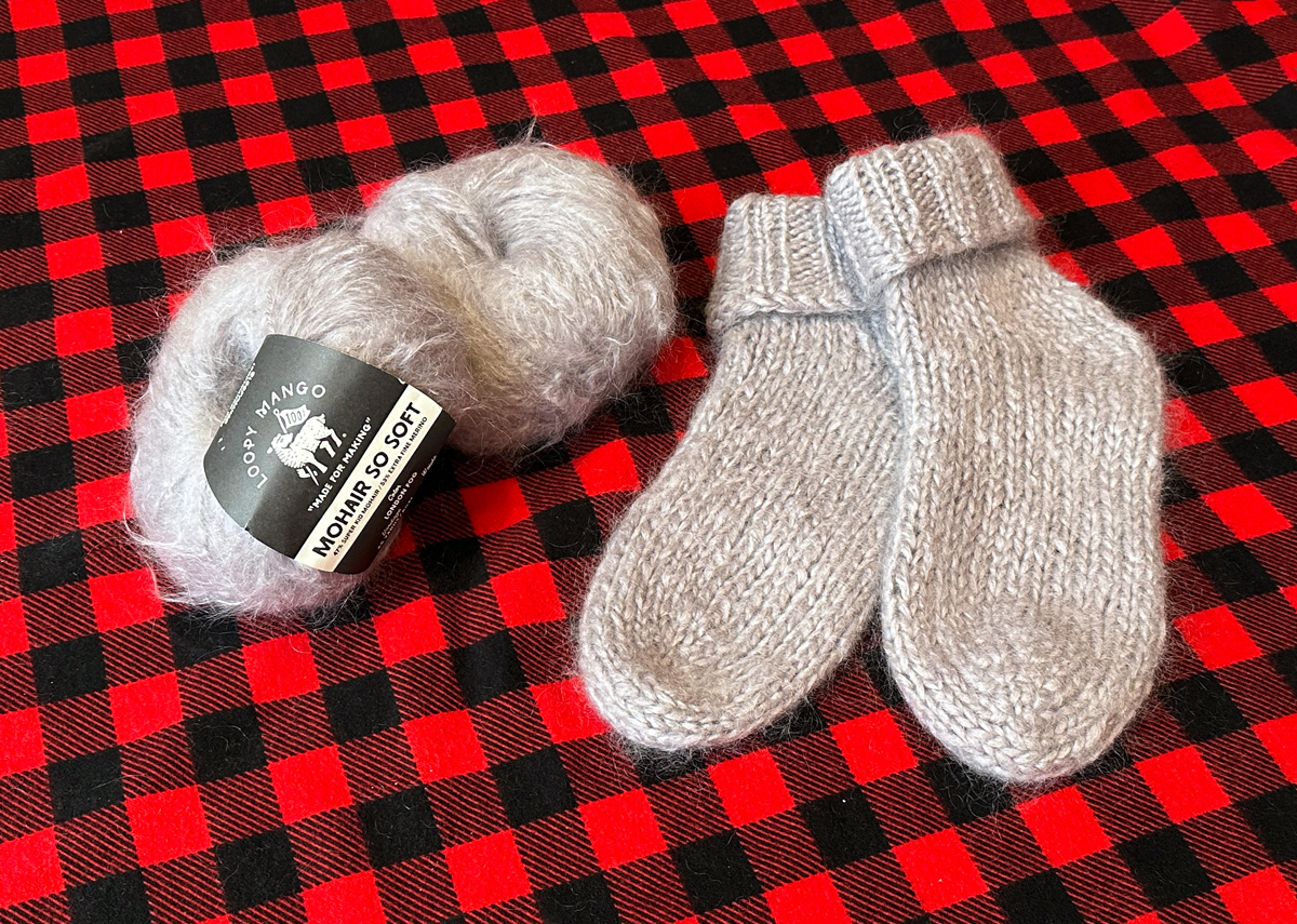Two fluffy balls of beige yarn along with a pair of fuzzy house socks knit in the same yarn lie on a red and black buffalo plaid cloth.
