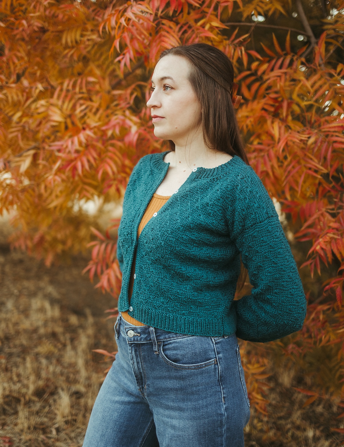 A white woman with long dark hair stands in front of flaming orange fall foliage wearing a Tunisian crochet cardigan in deep teal yarn.