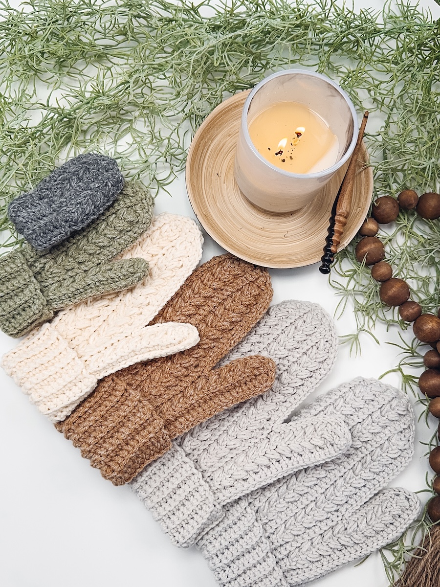 Six mittens in assorted sizes and colors lie next to holiday greenery and a lit candle