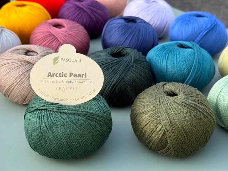 Shiny balls of yarn in assorted jewel tones sit on a light blue-green table.