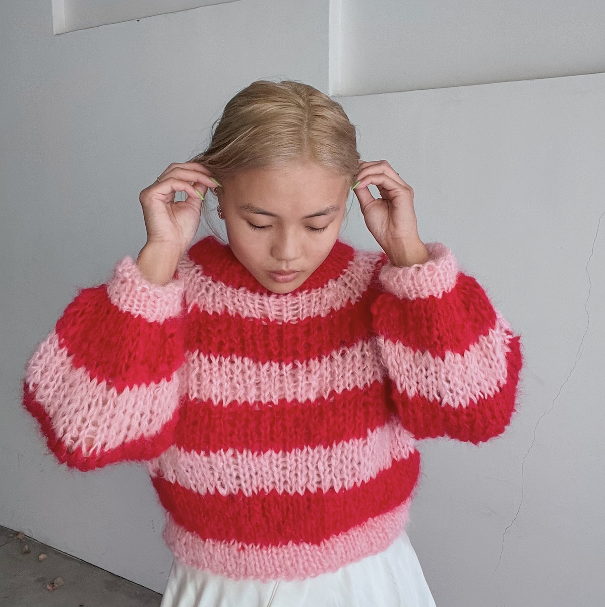 An Asian woman with blond hair and lime green nail polish models a fluffy sweater in bright pink and red stripes.