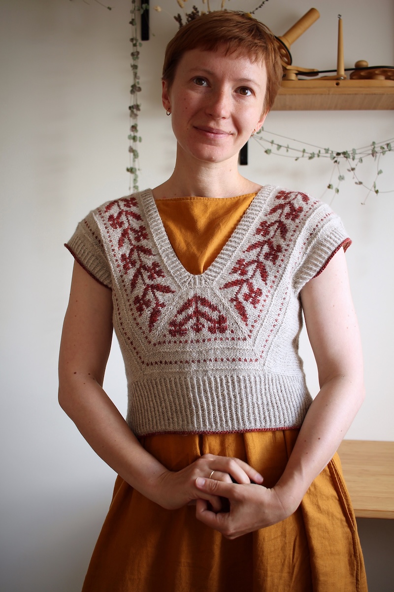 Designer Teti Lutsak, a white woman with short red hair, models a handknitted colorwork vest in beige and red over a warm yellow dress.