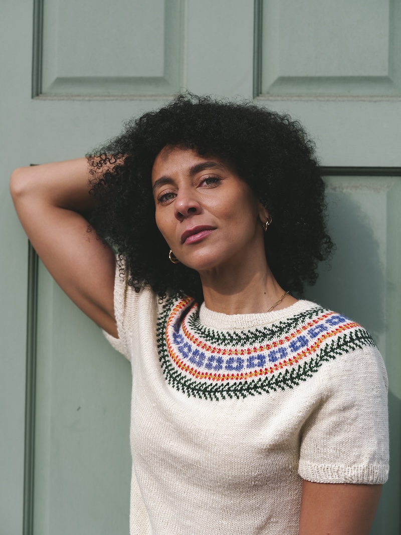 A brown skinned woman with curly dark hair models a hand knitted short sleeved sweater with a colorwork yoke.
