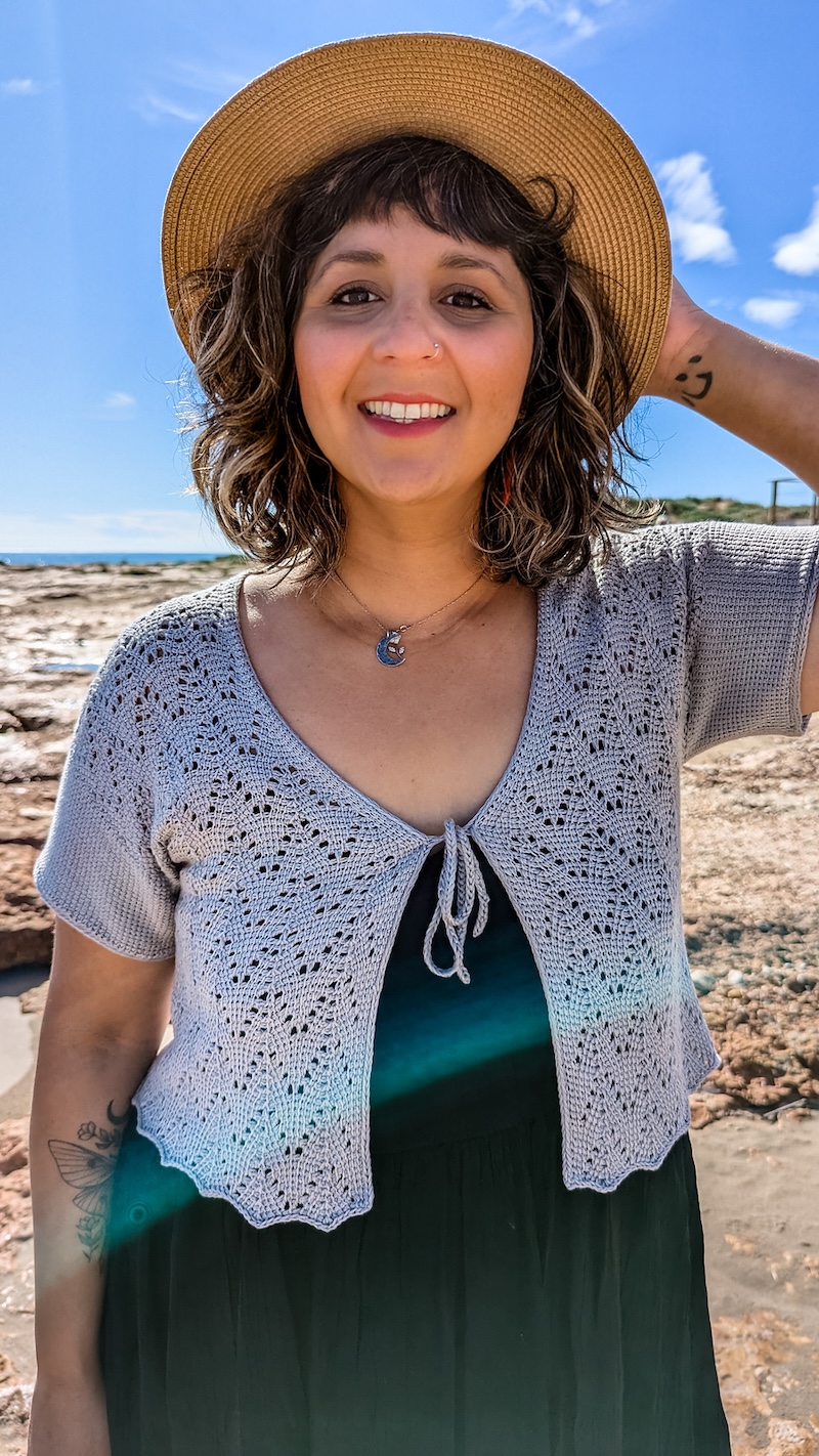 A smiling light-skinned woman with curly brown hair under a straw hat models a short-sleeved lace cardigan crocheted in pale blue yarn.