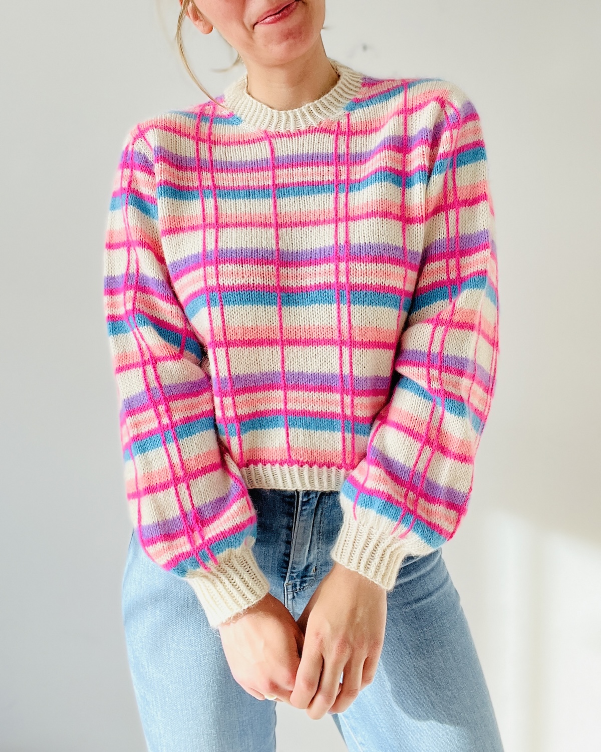 Closeup of a light-skinned person wearing a plaid pullover striped in bright pink, red, purple and blue on a white background.