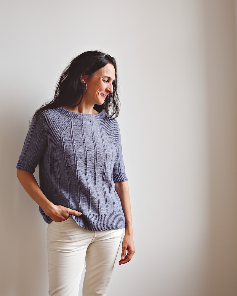 A white woman with long dark brown hair models a short sleeved hand knitted sweater in deep lavender yarn.
